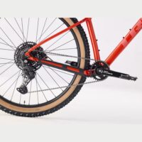 Red mountain bike rear drivetrain with cassette, derailleur, chain, and crankset visible on tan-wall tire.