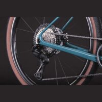 Rear bicycle drivetrain close-up: cassette, chain, and derailleur on a teal frame.