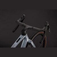 Close-up of a road bike's front end: drop handlebars, integrated brakes, gray speckled frame, and brown-tired wheel in a dark background