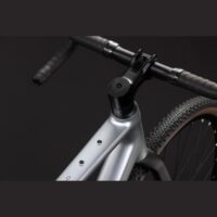 Close-up of a silver bicycle frame near the handlebar stem and black handlebars with a portion of the front tire visible on the right, set against a dark background.