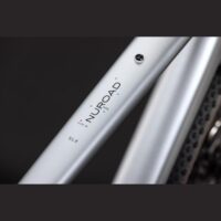 Close-up of a silver bicycle frame tube with the 'NUROAD' branding and small pink/blue marker dots, model SLX visible.